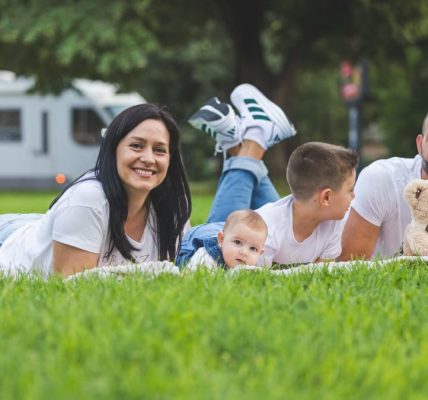 Happy family lying on grass with RV in the background, enjoying a sunny outdoor picnic.
